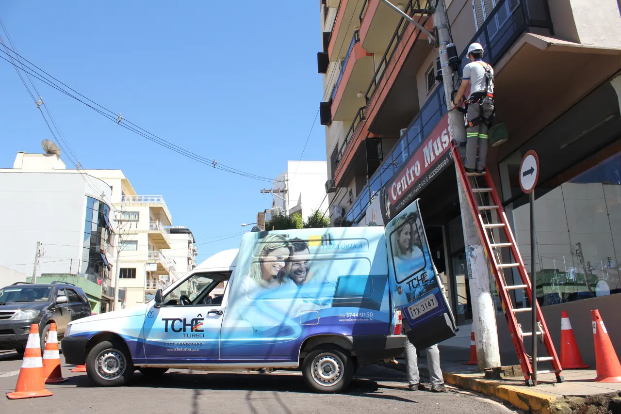 Técnico da Tchêturbo em uma escada, instalando fibra óptica na fachada de um prédio, com o carro da empresa estacionado na rua, em 2011