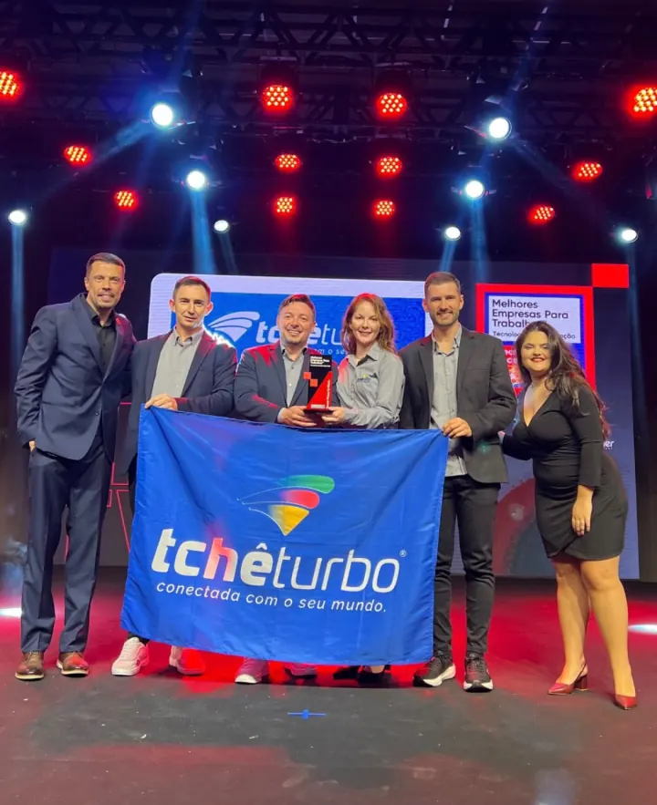 Equipe da Tchêturbo no palco com a bandeira da empresa e o troféu do GPTW de Tecnologia da Informação, em 2025